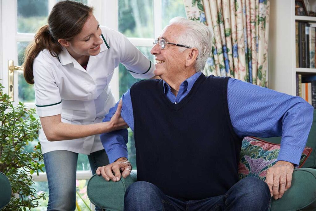 Two older adults chatting over tea at a small shared dining table in a sunny common room.