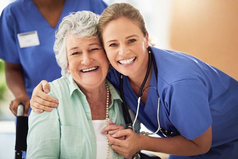 Two older adults, one wearing a rainbow pin, sitting together and smiling in a sunlit common area.